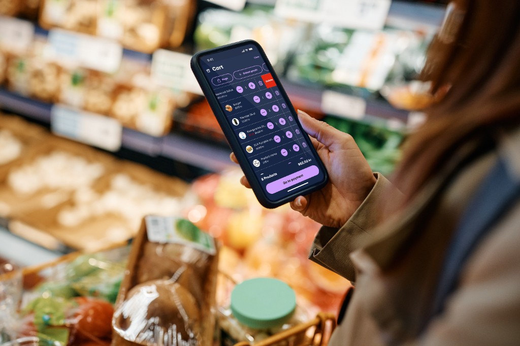 Shopper in a grocery aisle holding a phone showing the Focalpay cart — line items, swipe-to-delete, nine products and total in kr, and Go to payment on a dark UI