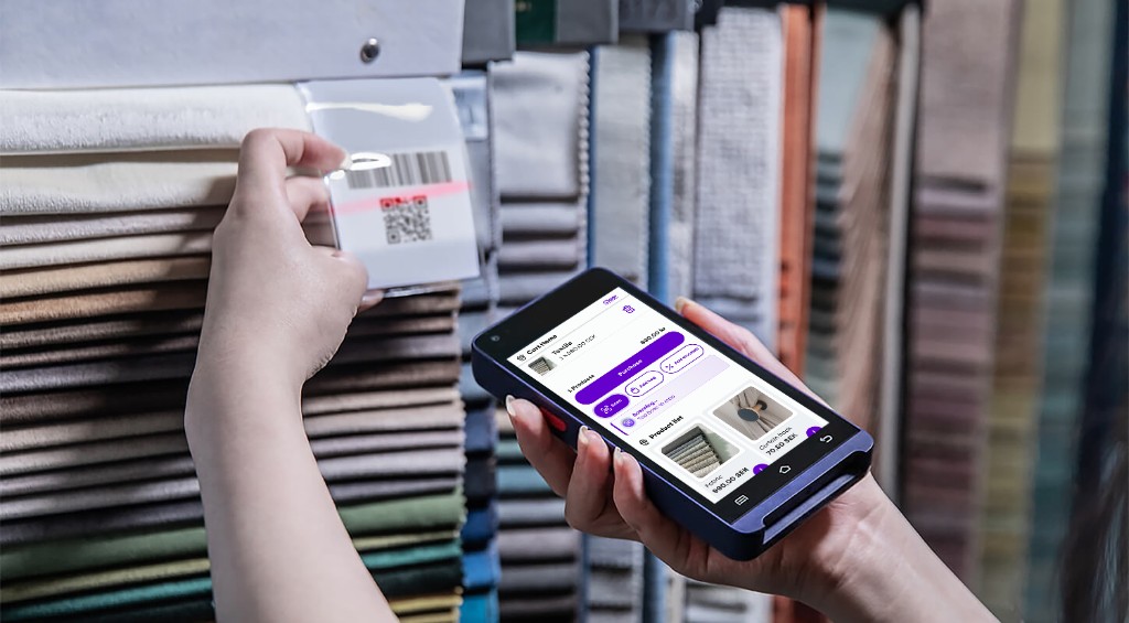 Cashier using a handheld mobile POS to scan a QR code on a fabric sample in a retail showroom, with cart and purchase actions on screen.